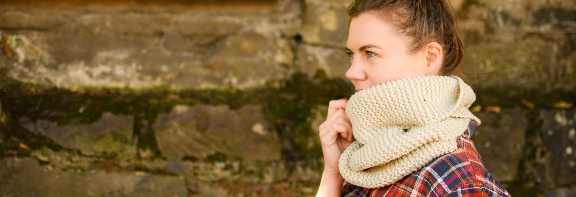 A white woman sits on the edge of a pavement with a wall behind her, she is smiling and wearing a cream coloured handknit scarf.